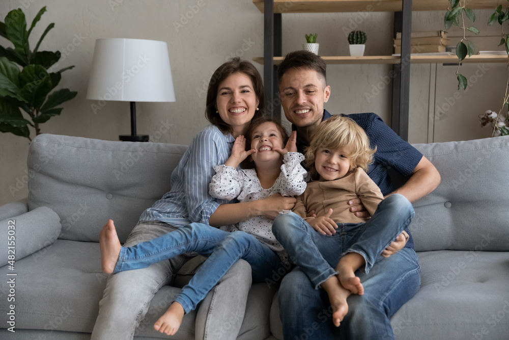 Smiling affectionate young couple parents holding on laps joyful little preschool son daughter kids, having fun spending leisure weekend time together at home, resting together on comfortable sofa.