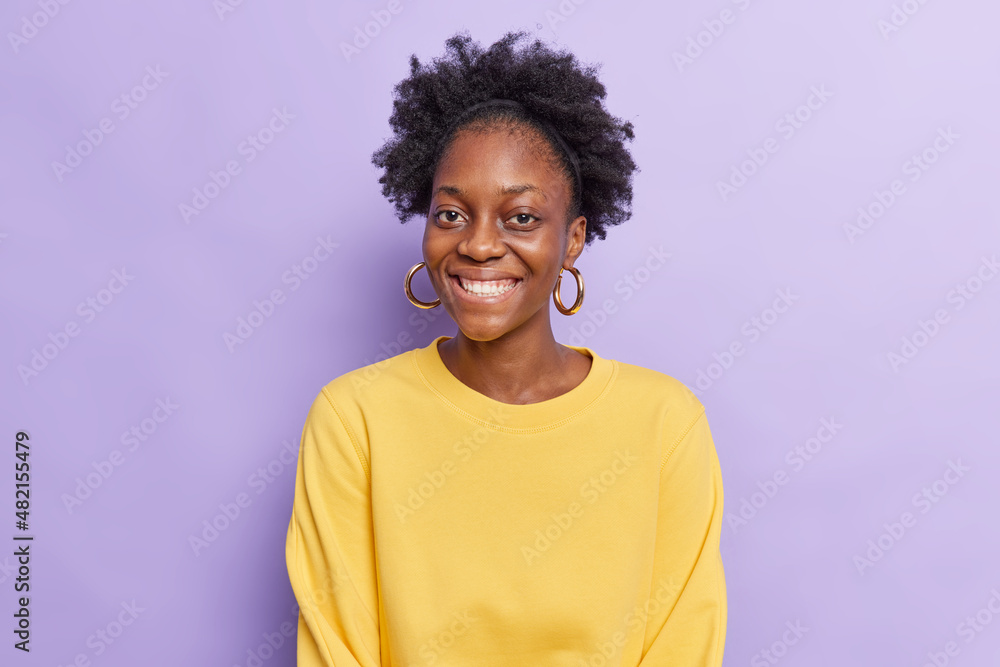 Portrait of cheerful dark skinned beautiful woman with curly hair smiles toothily wears casual yellow jumper has good mood isolated over purple background. Happy emotions and feelings concept Portrait of cheerful dark skinned beautiful woman with curly hair smiles toothily wears casual yellow jumper has good mood isolated over purple background. Happy emotions and feelings concept