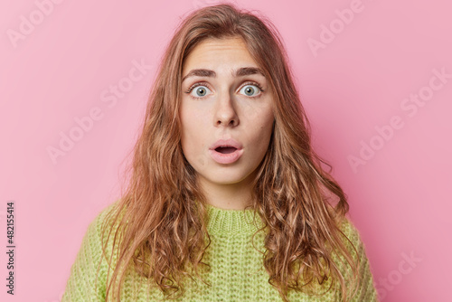 Shocked beautiful young long haired European woman stares bugged eyes keeps mouth opened reacts on amazing news wears green knitted sweater isolated over pink background. Human reactions concept © wayhome.studio 