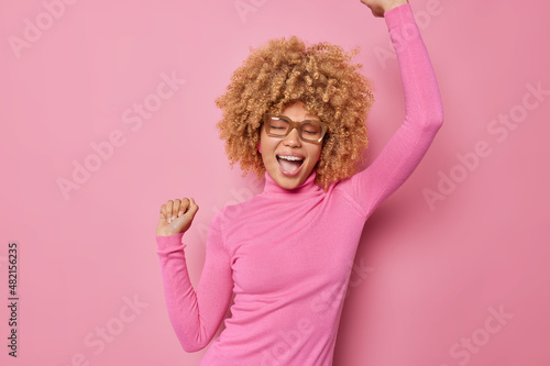 Image of carefree optimistic ambitious young woman does champion dance raises hands up celebrates something wears spectacles and turtleneck isolated over pink background triumps over success.