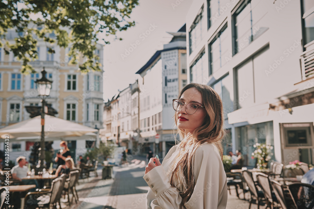 Fototapeta premium A slender girl with blond long hair in a white shirt and plaid skirt. Sunny weather girl walks around the city.