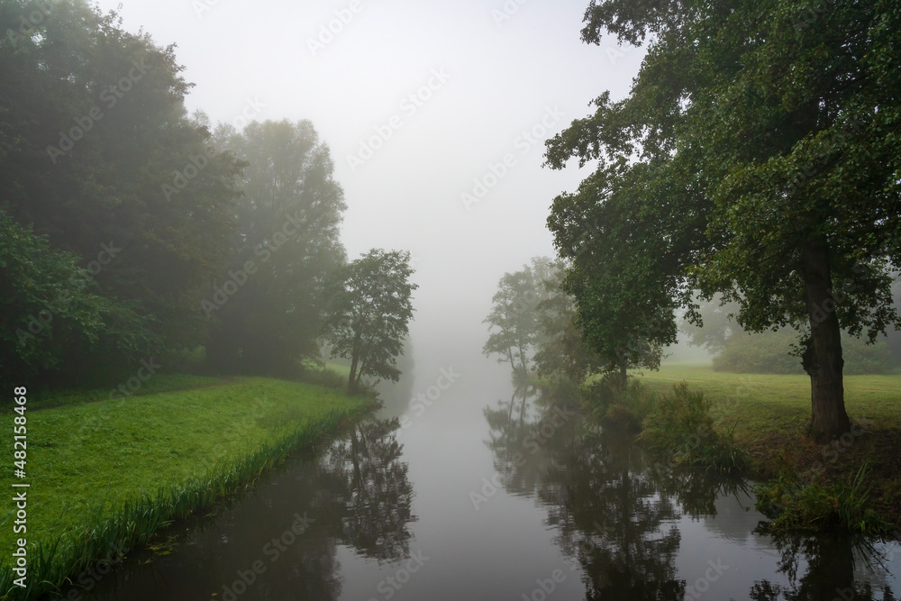The trees on either side of this ditch are beautifully reflected in the water of a ditch on a foggy morning in the Westerpark in Zoetermeer