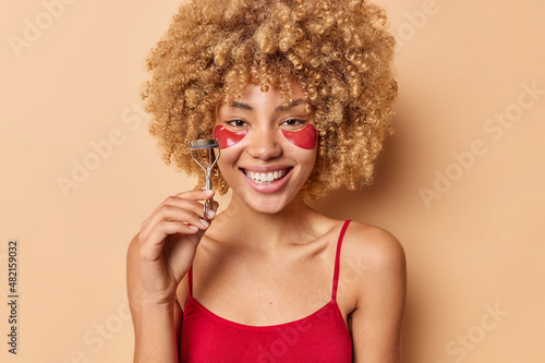 Positive beatiful woman with curly hair dressed in casual t shirt holds eyelashes curler going to apply makeup wears hydrogel patches under eyes being in good mood isolated over beige background