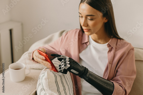 Selective focus of pretty attractive Caucasian brown-haired woman cyborg with black metal motorized artificial hand prosthesis using red smartphone sitting comfortably on couch, having rest