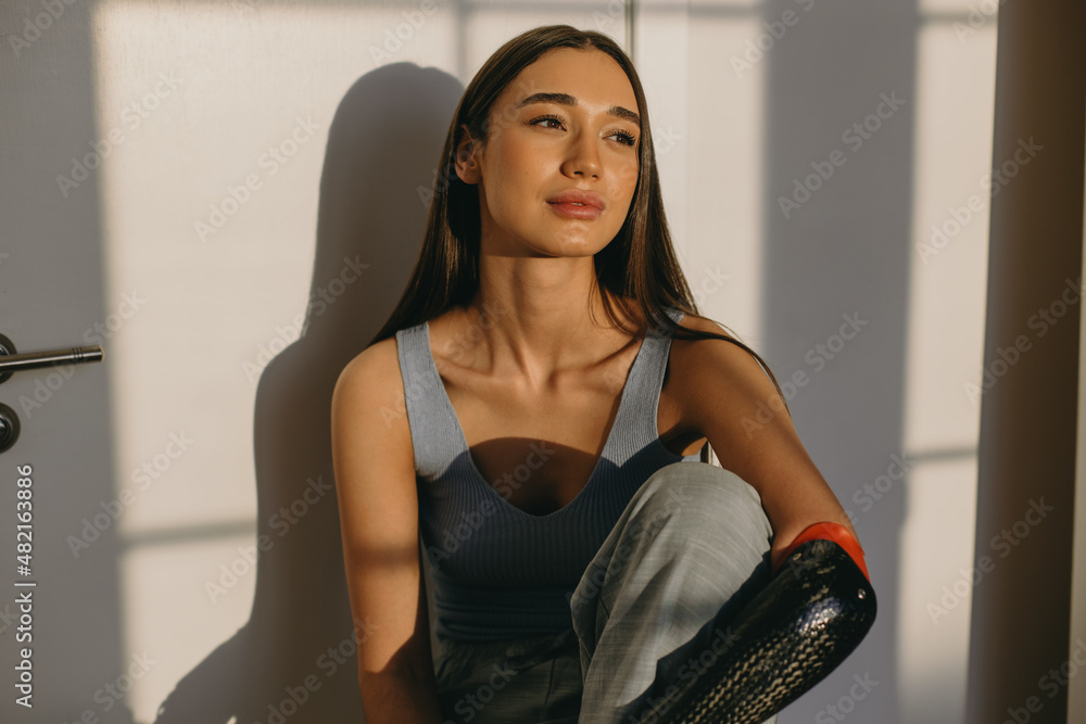 Fototapeta premium Closeup of charming lovely woman in blue top with bionic prosthesis on her left arm sitting against closed door, having pensive dreamy look, enjoying sunset, sun covering her cute perfect face