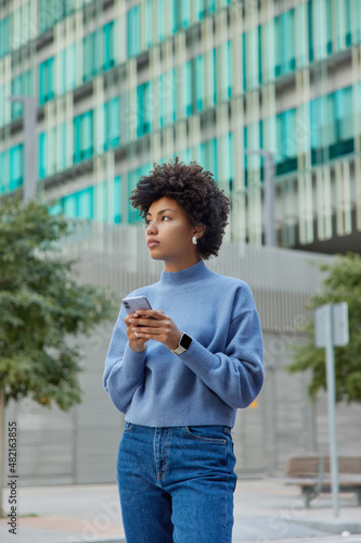 Outdoor shot of thoughtful beautiful woman wears casual blue jumper and jeans stands against urban setting uses mobile phone connected to wireless internet going to call taxi strolls in city