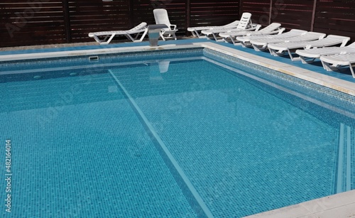 The pool with blue tiles is filled with water and empty white deck chairs.
