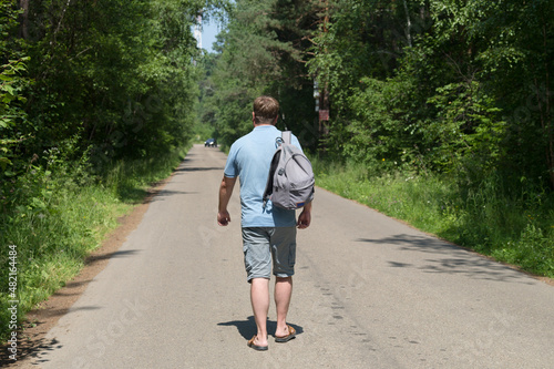 An adult male traveler with a backpack is walking alone on an empty road in the middle of the forest.