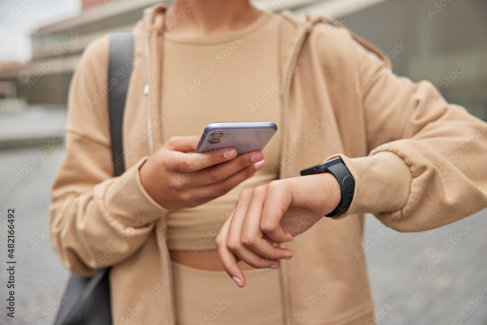 Cropped shot of unrecognizable woman holds modern mobile phone checks results of fitness traning dressed in sportswear carries fitness mat poses outdoors. People sport and technology concept Cropped shot of unrecognizable woman holds modern mobile phone checks results of fitness traning dressed in sportswear carries fitness mat poses outdoors. People sport and technology concept