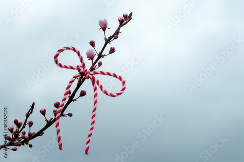 Hello spring background, March first holiday concept. Bow of red and white thread, martenitsa on blossoming branch of sakura or Japanese cherry against cloudy sky, copy space photo