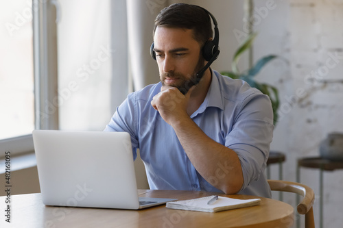 Millennial distant worker in headphones working from home, making video call to client on laptop, using computer at table, attending online virtual conference. Student watching learning webinar