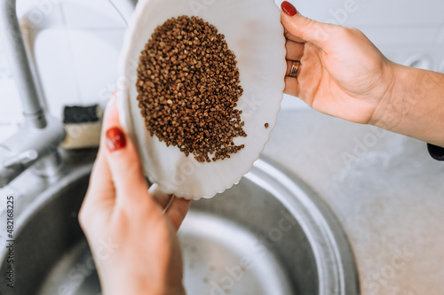 The girl, the cook, holds a plate in her hands and rinses buckwheat with water over the washbasin. photo