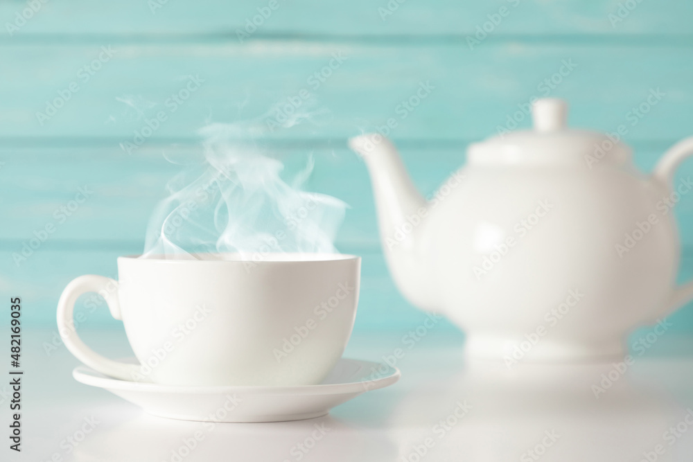 White cup with steam and teapot with herbal tea on blue wooden wall background White cup with steam and teapot with herbal tea on blue wooden wall background