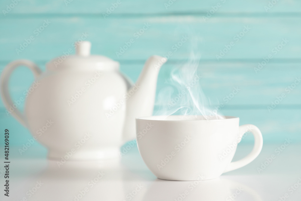 White cup with steam and teapot with herbal tea on blue wooden wall background White cup with steam and teapot with herbal tea on blue wooden wall background