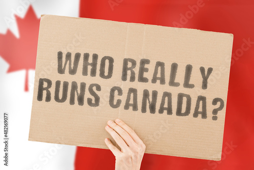 The quesiton " Who really runs Canada? " on a banner in men's hand with blurred Canadian flag on the background. Power. Authority. Government. Control. Politics. President. Outlaw. Hidden