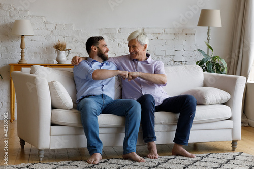 Happy mature grey haired dad congratulating adult son on success, giving friends fist bump gesture, support, praise, embracing grownup kid, laughing, sitting on couch in living room