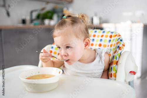 Cute baby girl toddler sitting in the high chair and eating her lunch soup at home kitchen.