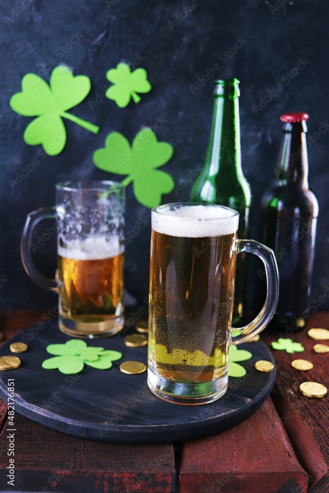 St. Patrick's Day, fresh beer in glass mugs and a bottle, gold coins, on a red wooden table, a green shamrock on a dark background, a party, a congratulation, a postcard St. Patrick's Day, fresh beer in glass mugs and a bottle, gold coins, on a red wooden table, a green shamrock on a dark background, a party, a congratulation, a postcard
