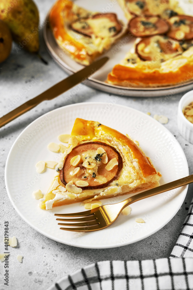 A slice of puff pastry pizza with pear, almonds, gorgonzola ricotta or blue cheese in a white plate on the kitchen table. Delicious layered pie with pear, dorblu, nuts on a light culinary background A slice of puff pastry pizza with pear, almonds, gorgonzola ricotta or blue cheese in a white plate on the kitchen table. Delicious layered pie with pear, dorblu, nuts on a light culinary background