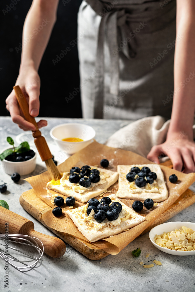 Raw puff pastry with cheese cream and blueberries. The process of making delicious sweet buns or mini pies. Greasing the edges of the baking dough with egg. Hands in the frame	