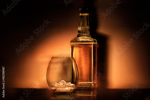 a bottle of alcohol rum whiskey or bourbon and a glass with ice on a dark orange background  retro still life