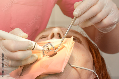 A patient with a cheek retractor with a green cofferdam in a dental clinic, a dentist in latex gloves examines her teeth with a boron and a mirror. photo
