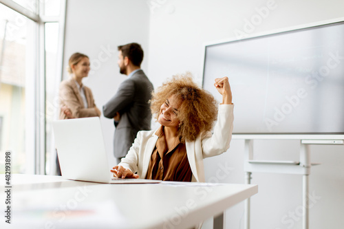 Young joyful businesswoman using laptop in the office with young people works behind her photo