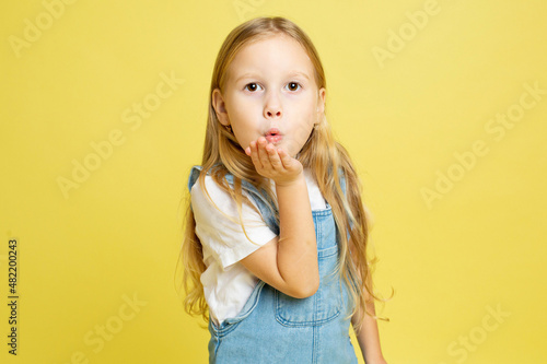 White european baby girl with blonde hair isolated on yellow blowing a kiss