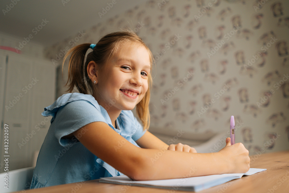Close-up of smiling elementary little child girl writing doing homework sitting at home table by window, looking at camera. Joyful primary kid studying alone making note with pen at bedroom. Close-up of smiling elementary little child girl writing doing homework sitting at home table by window, looking at camera. Joyful primary kid studying alone making note with pen at bedroom.