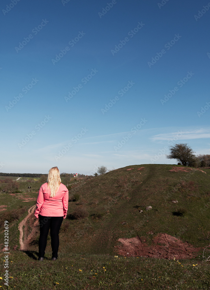 Woman with blonde hair standing and looking at view of hills, fields and open landscape on a summer day. Woman wearing pink windbreaker jacket. Photo taken at Brösarps Backar in Skåne, Sweden.