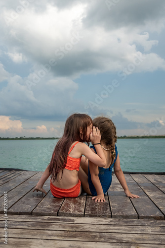 Small girls sitting on the lake wooden pier, enjoying summer vacation, talking about secrets, having fun. 