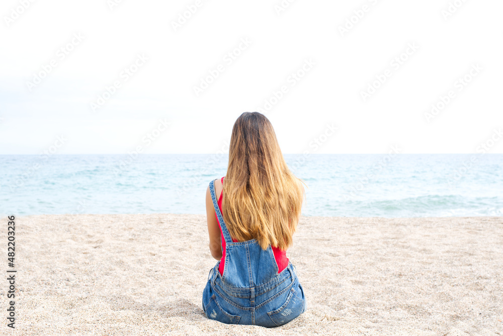 Lonely girl with long brown hair and jeans sitting on the sand of the beach looking at the mediterranean sea. Back view.
