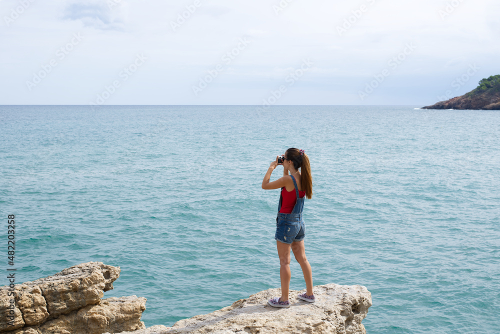 Girl with jean shorts and sneakers standing in a rock photographing the mediterranean sea during her vacations. Summer leisure and hobbies.
