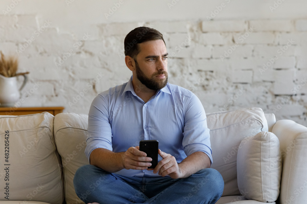 Serious business man using online app on mobile phone on couch at home, looking away. Smartphone user lost in thoughts, thinking over text message, problem, feeling doubt, worried, uncertain Serious business man using online app on mobile phone on couch at home, looking away. Smartphone user lost in thoughts, thinking over text message, problem, feeling doubt, worried, uncertain