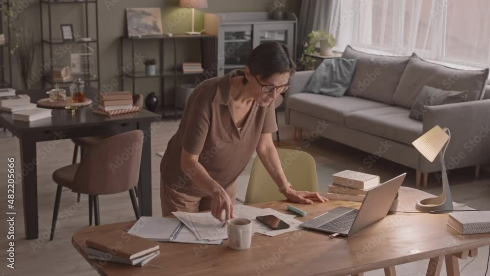 Medium slowmo shot of Caucasian mid-adult woman standing by desk with piles of notes and study materials watching educational video lesson on laptop studying from home Medium slowmo shot of Caucasian mid-adult woman standing by desk with piles of notes and study materials watching educational video lesson on laptop studying from home