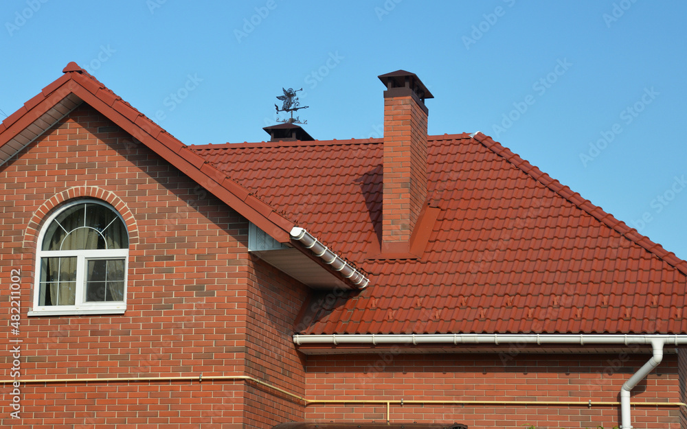 A ceramic, clay roof of a brick house with a chimney, flashing, attic window, a weather vane, fascia, and soffit, and a roof gutter with a downpipe.