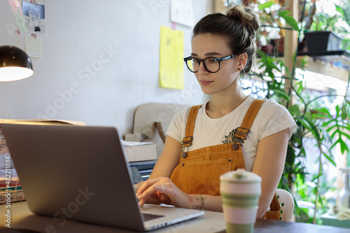 Female flower shop owner taking online customer orders, young woman florist or gardener working on computer ordering gardening products via internet, sitting at cozy workplace at home, selective focus