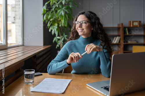 Positive happy female employee resting at workplace, looking away from computer screen. Relaxed woman office worker taking break to refresh mind and prevent stress during workday. Employee wellbeing photo