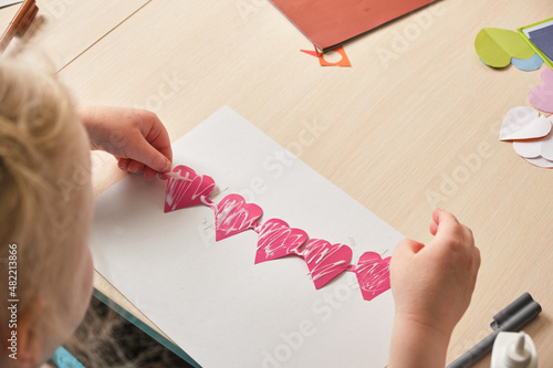 preschooler girl in a blue t-shirt makes an applique sitting at the table, hearts cut out of colored bougue for a card photo