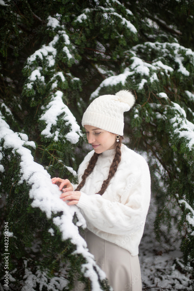 Young woman with white skin and long pigtails near spruce covered with snow dressed in warm white clothes