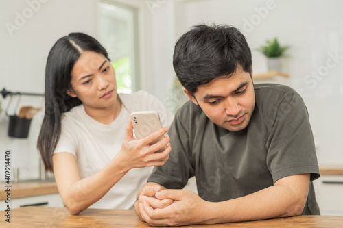 Infidelity, suspicion asian young couple love fight relationship, wife holding cellphone, smartphone cheating on phone, scolding husband about mistrust, distrust and jealousy when sitting at home.