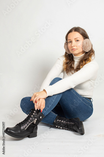 Young woman in warm white jumper with fur earmuffs isolated on white sitting on the floor with crossed legs in jeans