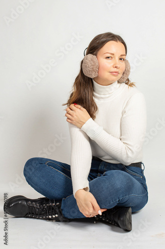 Young woman in warm white jumper with fur earmuffs isolated on white sitting on the floor with crossed legs in jeans