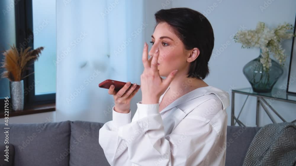 Portrait of young caucasian woman standing in apartment livivng room. Using smart phone and recording voice message. Smiling attractive female using modern internet technologies close up. 