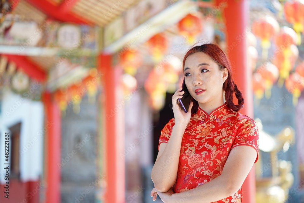 Portrait beautiful Asian young woman wearing red traditional Chinese cheongsam decoration and using smartphone for Chinese New Year Festival at Chinese shrine