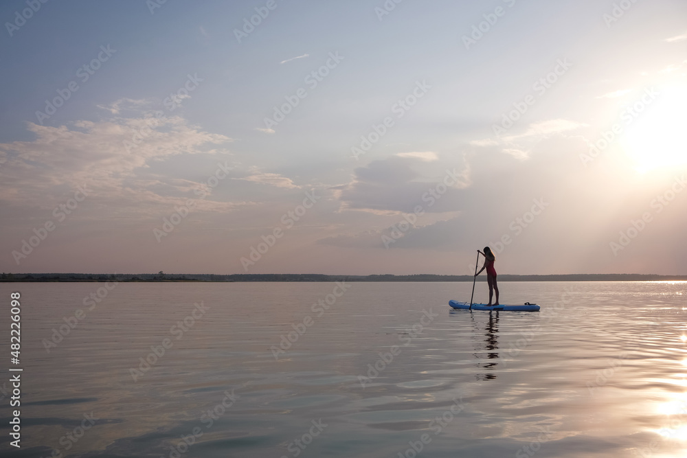 Silhouette of a beautiful woman swimming on big lake staying Paddle SUP Board at sunset background.