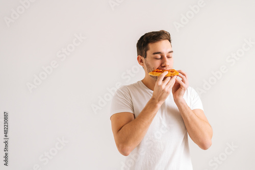 Portrait of happy young man with enjoying eating delicious slice of pizza, with closed eyes from pleasure on white isolated background. Studio shot of hungry handsome male student eating tasty food.