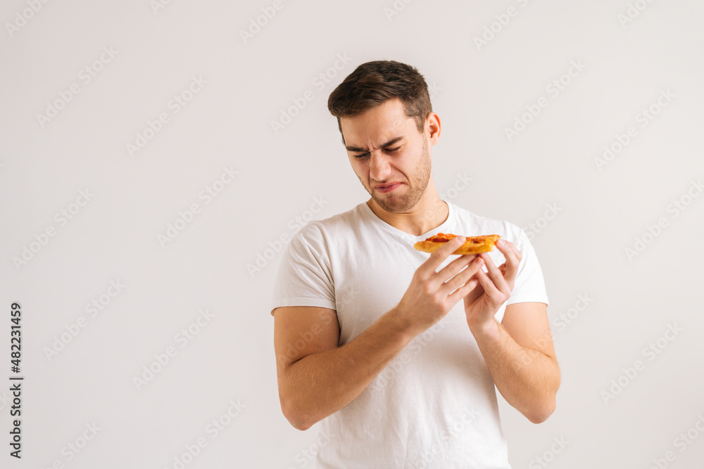 Portrait of frustrated sad young man holding piece of pizza with awful smell, smelling tainted unappetizing food, dissatisfied with food quality on white isolated background in studio. Portrait of frustrated sad young man holding piece of pizza with awful smell, smelling tainted unappetizing food, dissatisfied with food quality on white isolated background in studio.