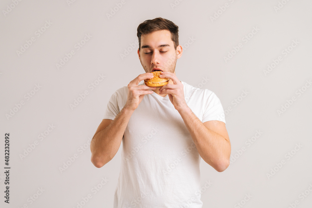 Portrait of handsome young man bitting eating delicious burger with closed eyes on white isolated background. Studio shot of happy hungry male holding tasty unhealthy hamburger indoors. Portrait of handsome young man bitting eating delicious burger with closed eyes on white isolated background. Studio shot of happy hungry male holding tasty unhealthy hamburger indoors.