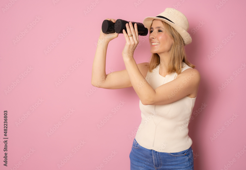 Young woman wearing straw hat and looking in the distance with binoculars on isolated pink background Young woman wearing straw hat and looking in the distance with binoculars on isolated pink background